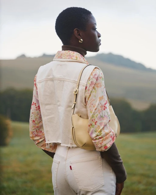 Woman in casual fashion stands in an outdoor setting with rolling hills; she wears light-wash jeans and vest, a floral top, and a purse.