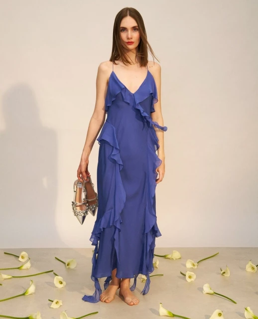 Full-length shot of a woman in a blue dress, holding embellished heels, surrounded by white flowers in a bright, neutral studio setting.