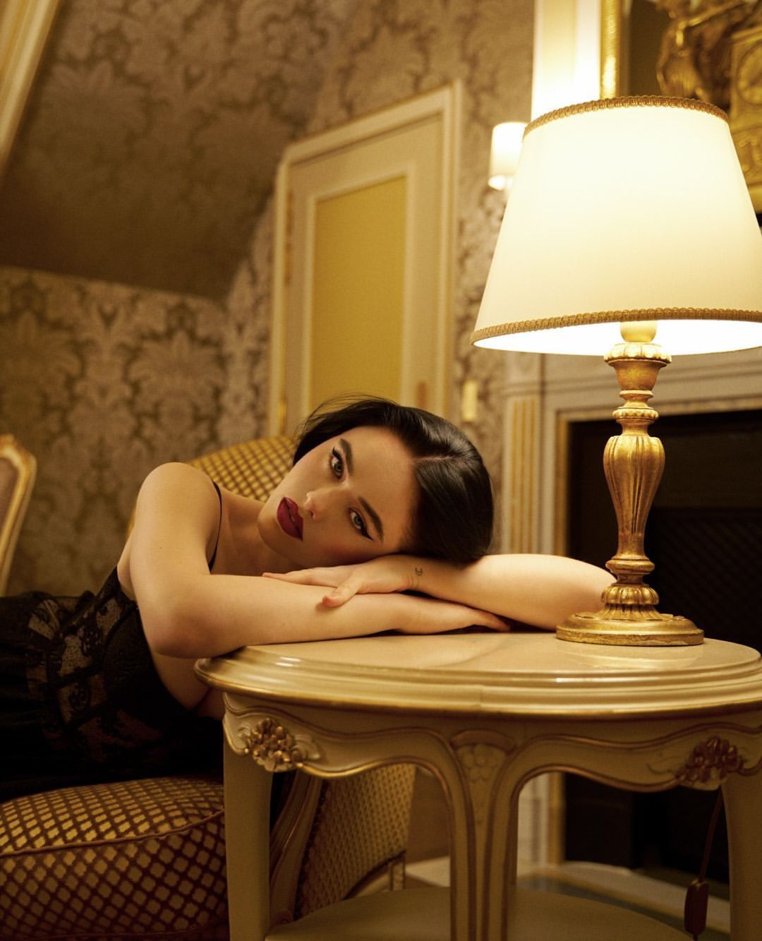 Adult woman with dark hair lies on a table in an ornate room. A lamp sits on the table near her head. Interior design.