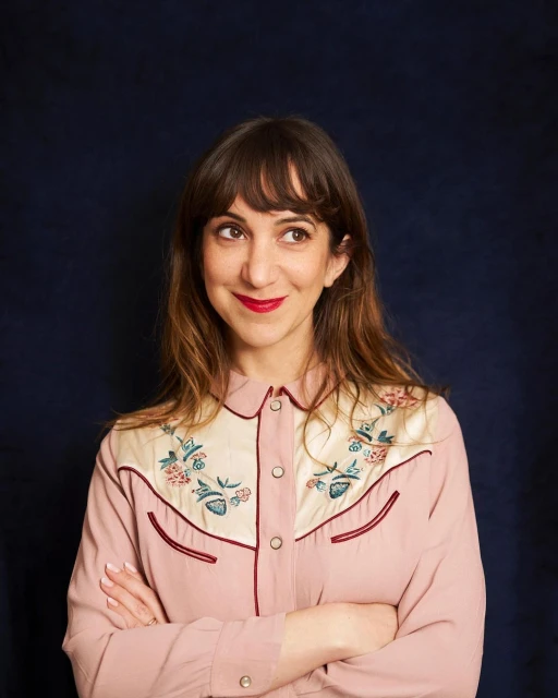 Portrait of a smiling adult woman with medium-length brown hair, wearing a light pink western-style shirt with floral embroidery, indoors.
