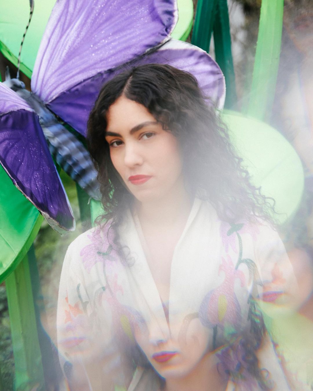 Portrait of a young woman with dark curly hair and red lipstick, wearing a floral kimono, standing in front of a purple butterfly prop.