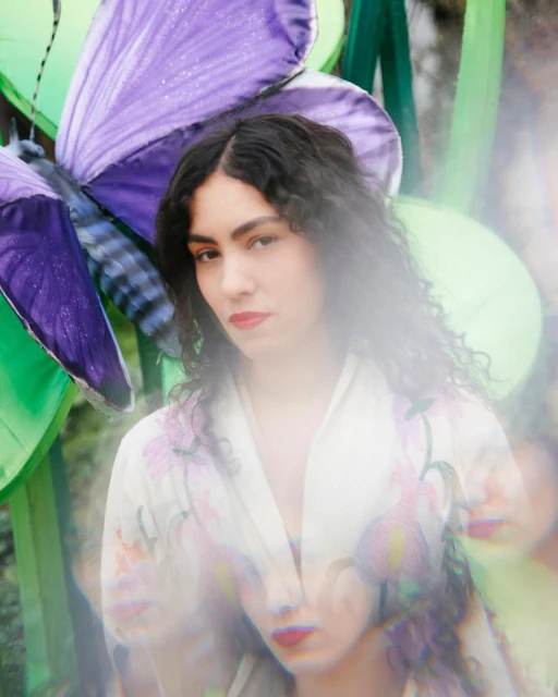 Portrait of a young woman with dark curly hair and red lipstick, wearing a floral kimono, standing in front of a purple butterfly prop.