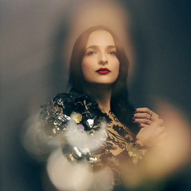 Portrait of a woman with dark hair and red lipstick wearing a floral-embellished black top against a grey background with bokeh effects.
