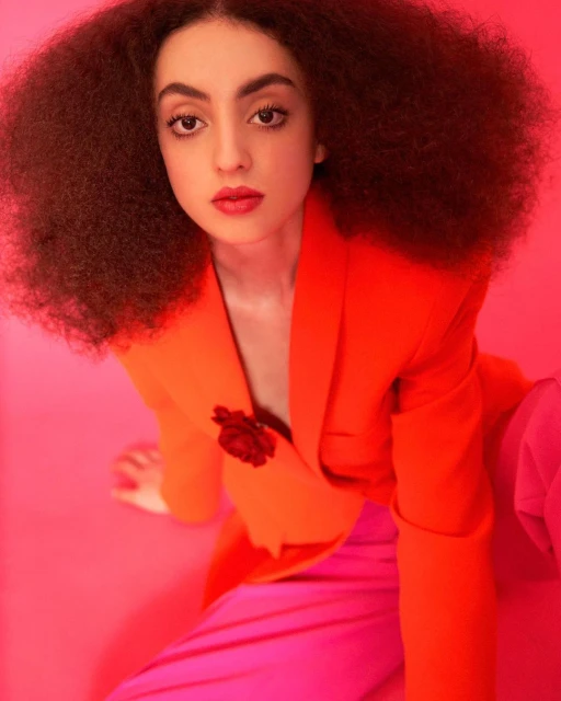 A young woman with curly dark hair and red lipstick poses indoors wearing a vibrant orange blazer and pink pants against a bright pink background.