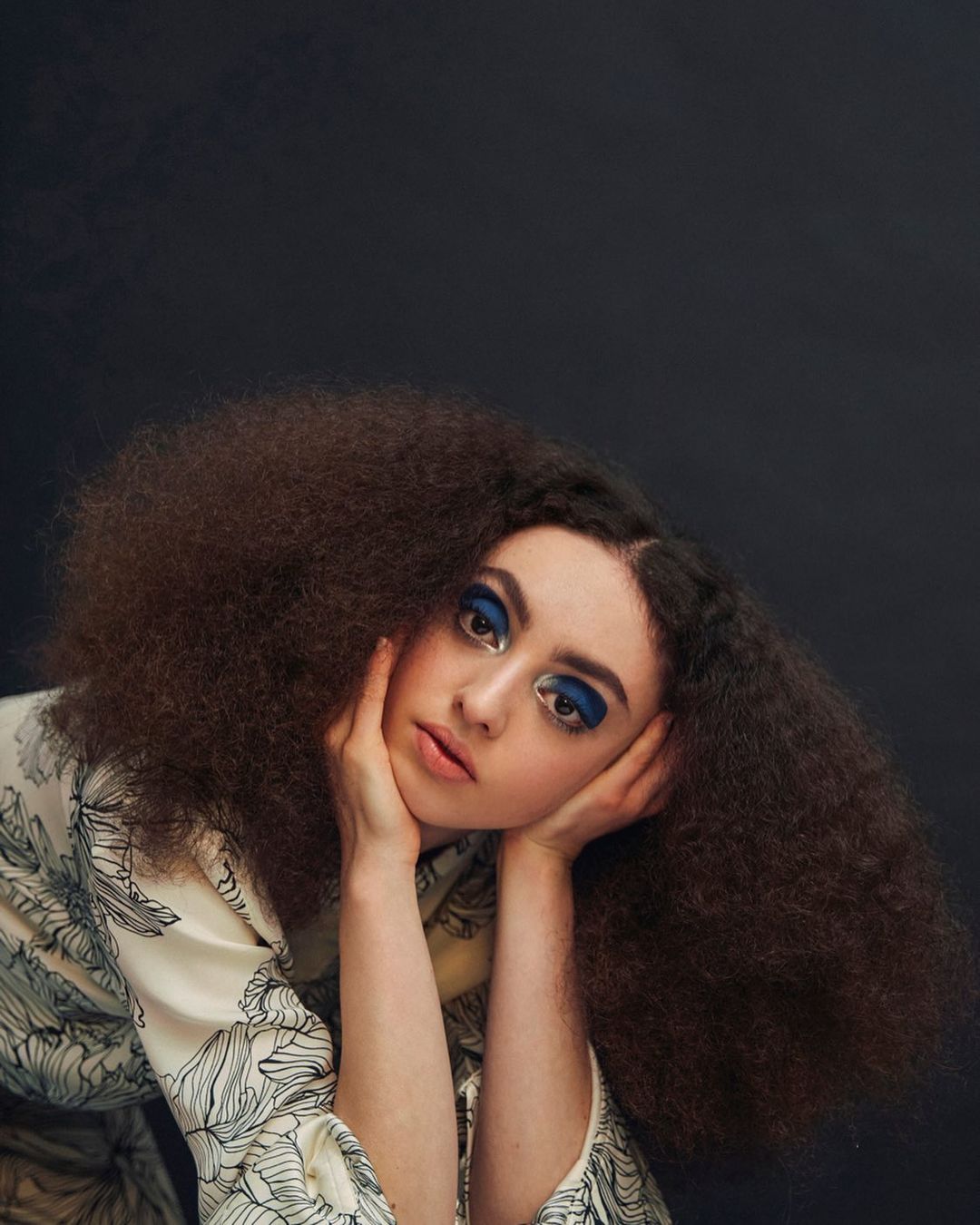 Close-up portrait of an adult female with afro hairstyle, bold blue eye makeup, and floral print top against a dark background.