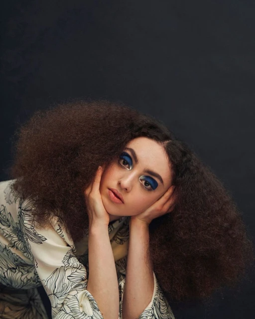Close-up portrait of an adult female with afro hairstyle, bold blue eye makeup, and floral print top against a dark background.