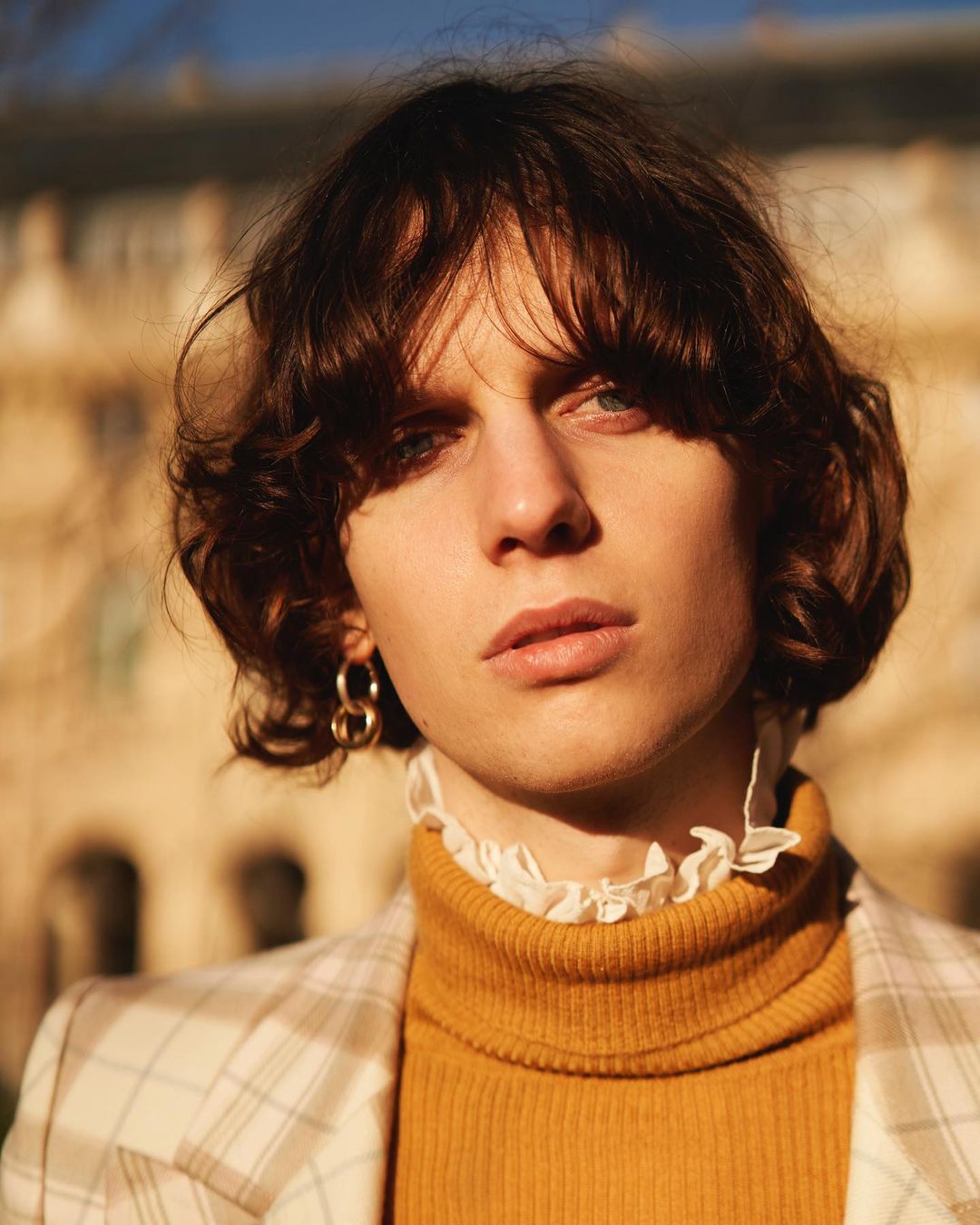 Portrait of a person with dark hair wearing a yellow turtleneck and plaid jacket against an outdoor architectural background.