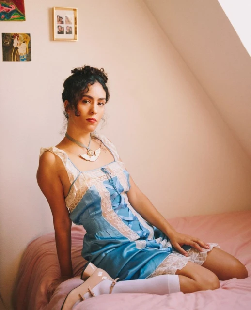 Woman with curly hair wearing a blue slip dress and Mary Jane shoes sitting on a pink bed in an indoor setting with art on the wall.
