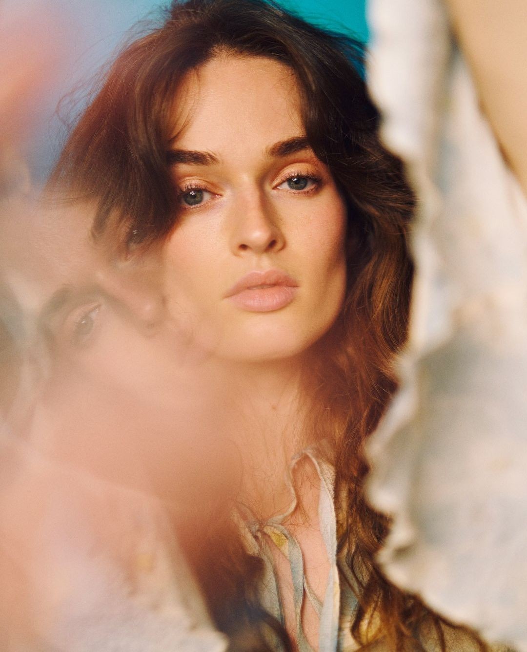 Close-up portrait of a woman with brown hair and light eyes, wearing a light-colored top against a teal and white background.