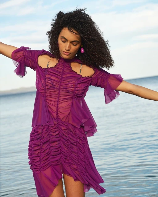 A curly-haired woman in a purple dress stands on the beach with arms outstretched toward the ocean and sky in a fashion-style shot.