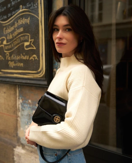 Brunette woman in cream sweater with black purse in urban setting, standing near building with French text.