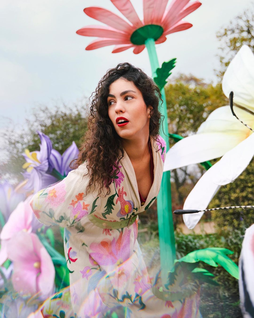 Portrait of a woman with curly hair and floral dress standing among colorful, oversized flowers in an outdoor garden setting on a cloudy day.