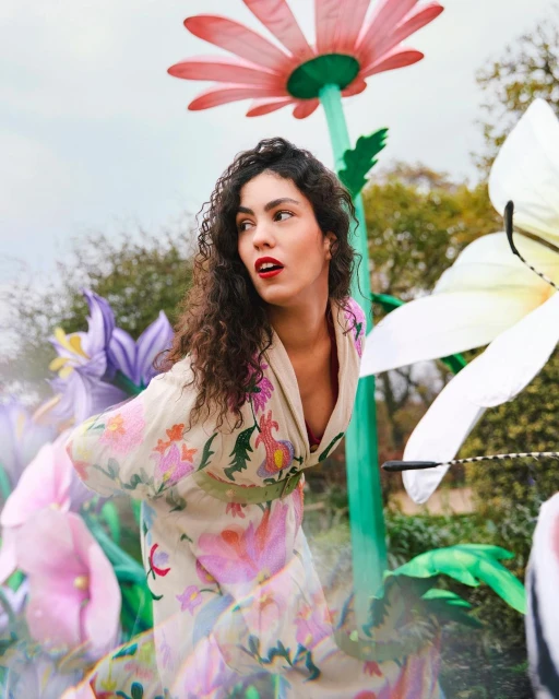 Portrait of a woman with curly hair and floral dress standing among colorful, oversized flowers in an outdoor garden setting on a cloudy day.