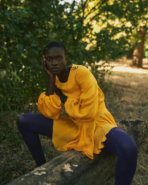 A young Black woman in a yellow dress poses on a log in a sun-dappled forest. Fashion editorial portrait. Autumnal tones.