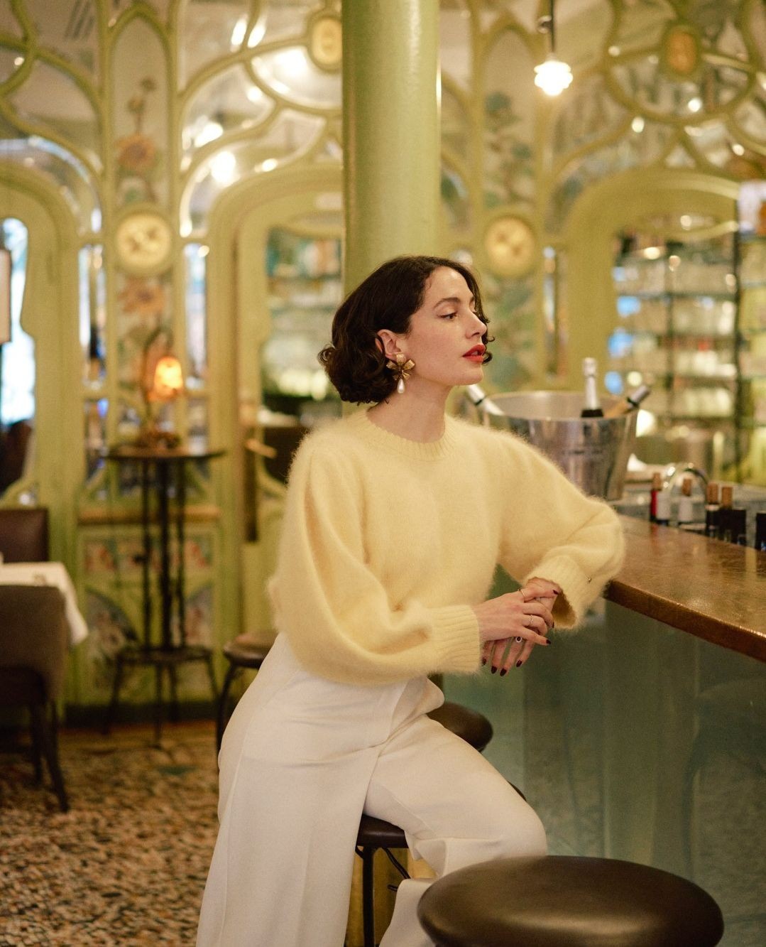 Elegant woman poses in a restaurant wearing a yellow sweater and white pants, sitting on a stool with ornamental details in background.