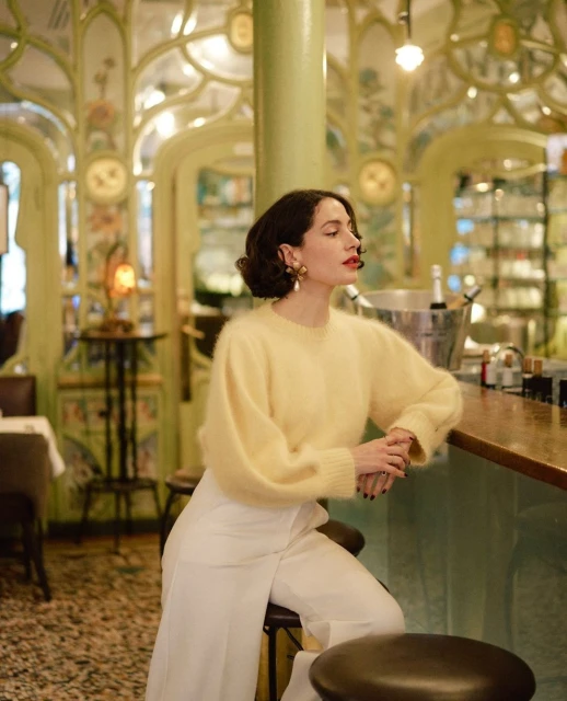 Elegant woman poses in a restaurant wearing a yellow sweater and white pants, sitting on a stool with ornamental details in background.