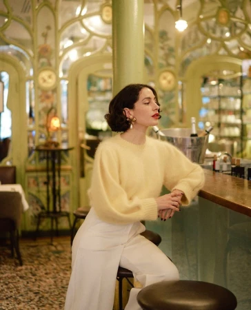 Elegant woman poses in a restaurant wearing a yellow sweater and white pants, sitting on a stool with ornamental details in background.