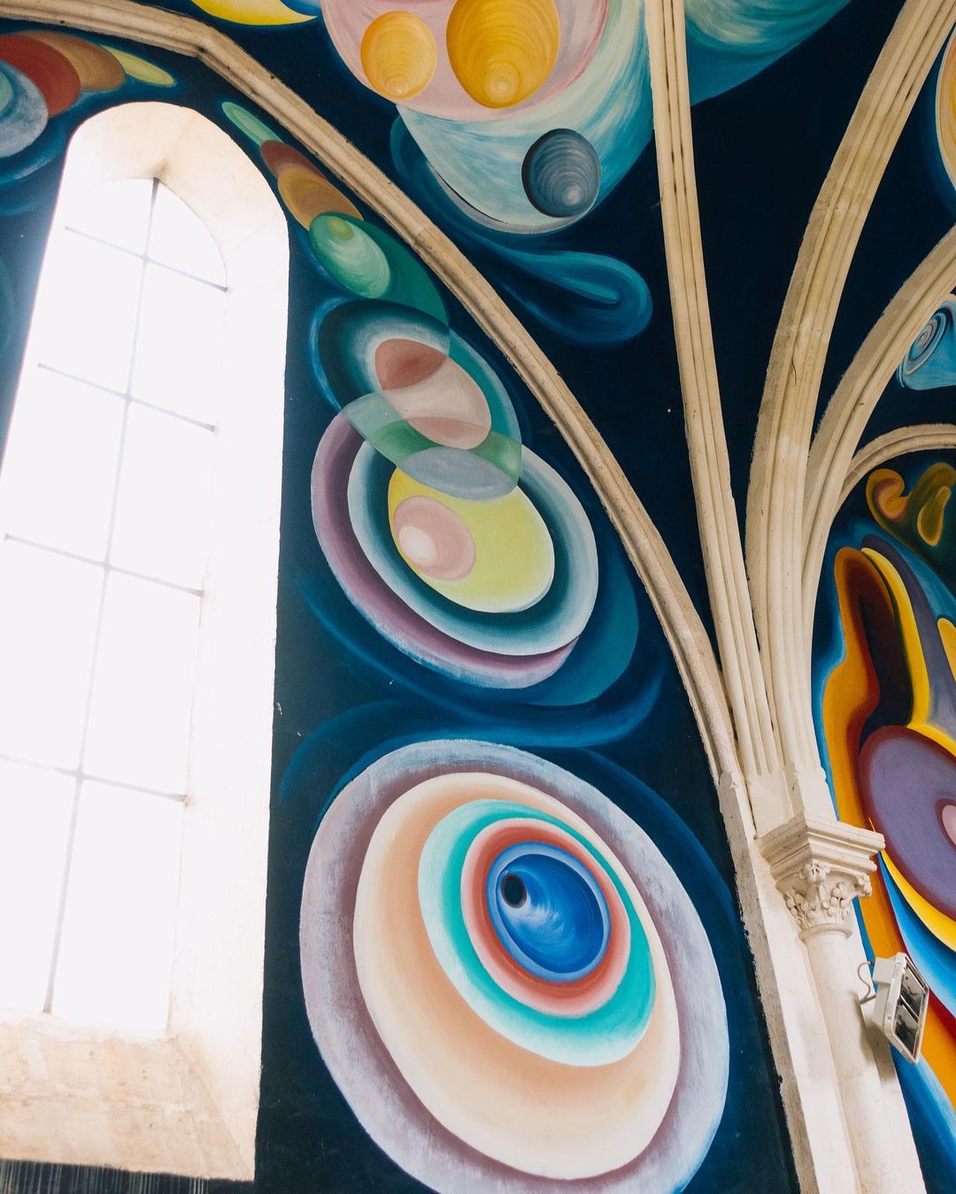 Interior view of a church ceiling featuring a vibrant mural with colorful circles and shapes and a bright white window in the background.