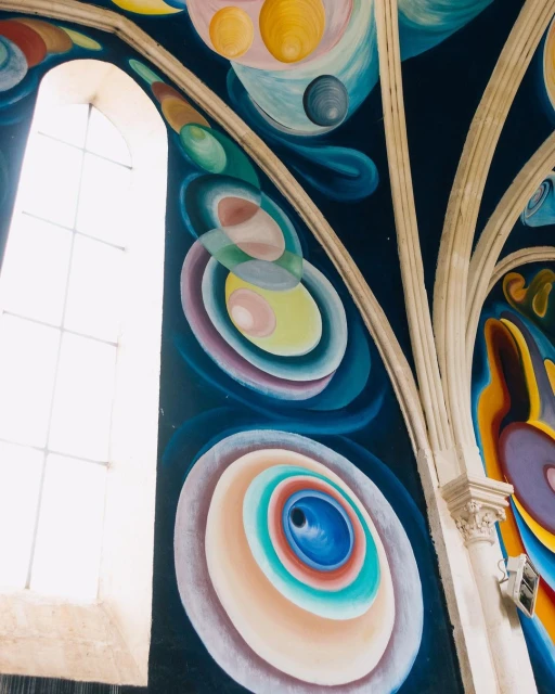 Interior view of a church ceiling featuring a vibrant mural with colorful circles and shapes and a bright white window in the background.