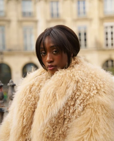 Young woman with dark hair poses in a cream shaggy coat, set against an urban backdrop of buildings with arched windows and ornate architecture.