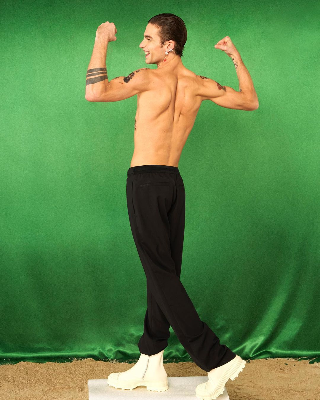 Athletic man with tattoos flexes muscles in a studio, wearing black pants and white boots, against a green backdrop.