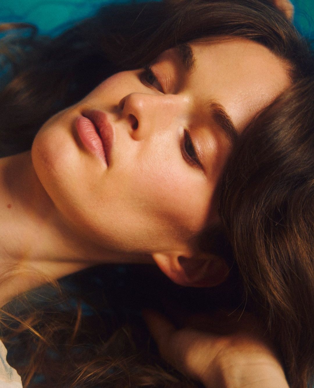 Close-up portrait of a serene woman with closed eyes, showcasing soft, warm skin tones against a blurred turquoise background. Relaxed expression.