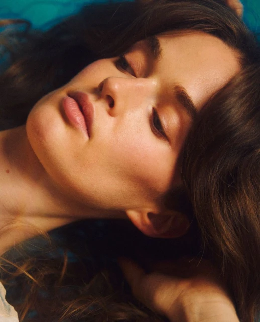 Close-up portrait of a serene woman with closed eyes, showcasing soft, warm skin tones against a blurred turquoise background. Relaxed expression.