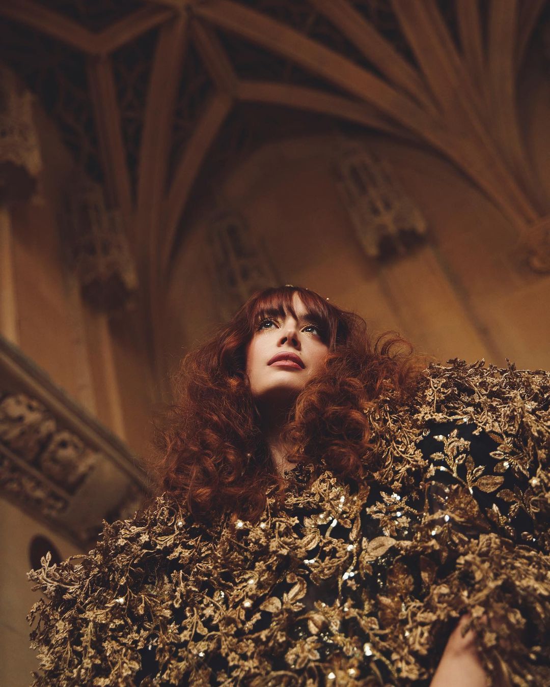 Woman with curly red hair wears a gown embellished with metallic flowers in a cathedral with ornate arches and neutral stone walls.