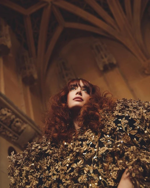 Woman with curly red hair wears a gown embellished with metallic flowers in a cathedral with ornate arches and neutral stone walls.