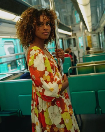 Adult female with curly hair in a vibrant floral print dress poses inside a passenger train car with green seats and silver poles.