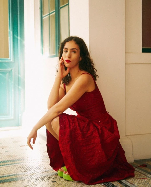 Woman in red dress poses in a squatting position indoors with bright green platform shoes in a medium shot looking at the camera.