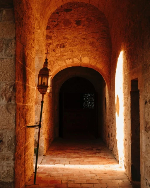Exterior shot of a stone hallway with arches and a decorative lamp illuminating a brick-paved floor and creating an atmospheric mood.