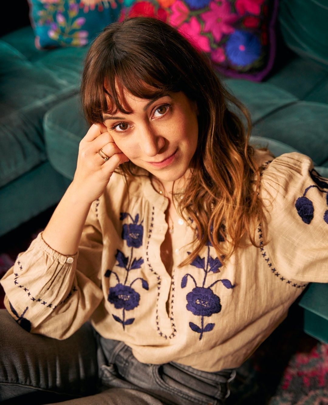 A brunette woman with bangs poses indoors, wearing an embroidered blouse, with a teal couch and floral pillows in soft, diffused light.