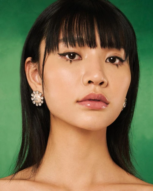 Close-up portrait of a young adult Asian woman with dark hair and pearl earrings against a green background in a beauty shot.