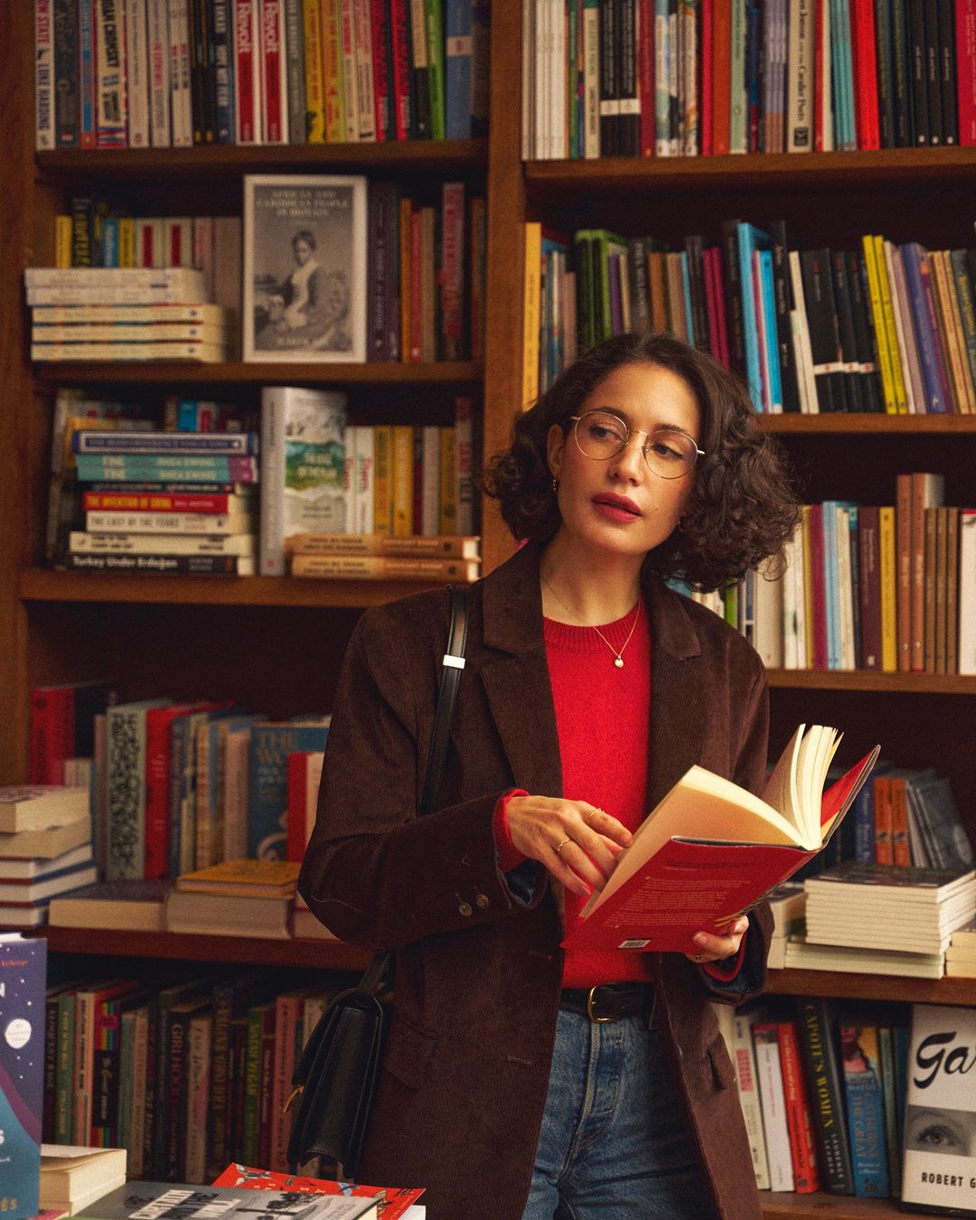 Woman with curly hair in a red sweater and brown corduroy jacket reading a book in a library with bookshelves in background.