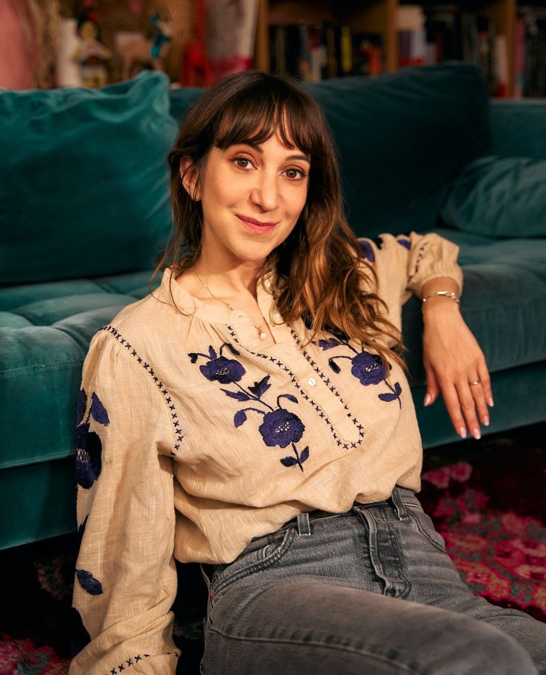 Portrait of a woman with long brown hair wearing an embroidered linen shirt and jeans, sitting on a turquoise sofa in a home interior.