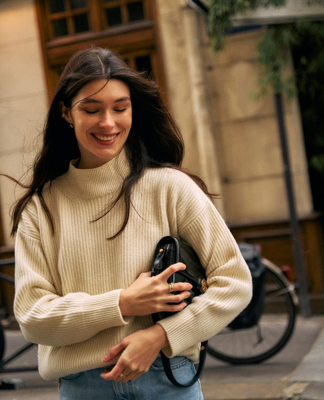Smiling woman in a cream sweater and jeans walks on an urban sidewalk carrying a black purse in daylight, with a bicycle in the background.
