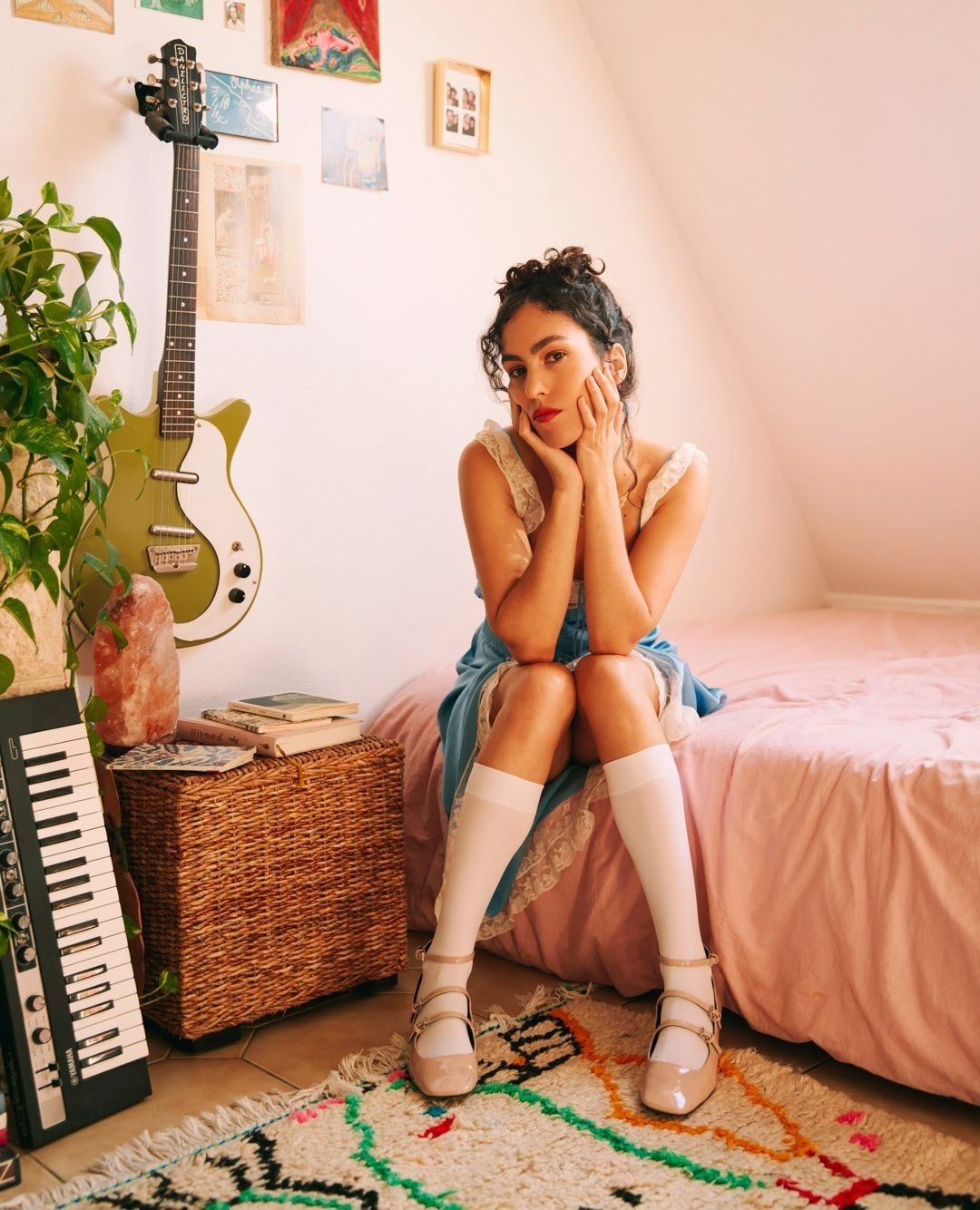 Young woman with curly hair, wearing a blue dress, sitting on a pink bed with an electric guitar and keyboard in a room setting.