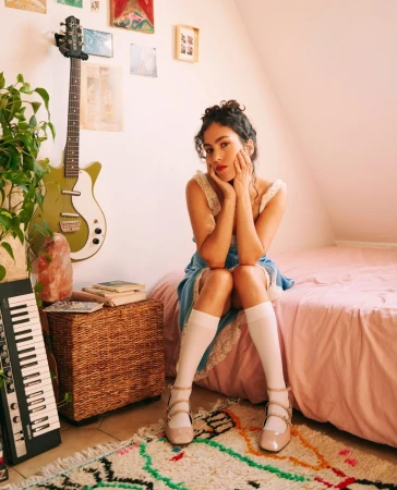 Young woman with curly hair, wearing a blue dress, sitting on a pink bed with an electric guitar and keyboard in a room setting.