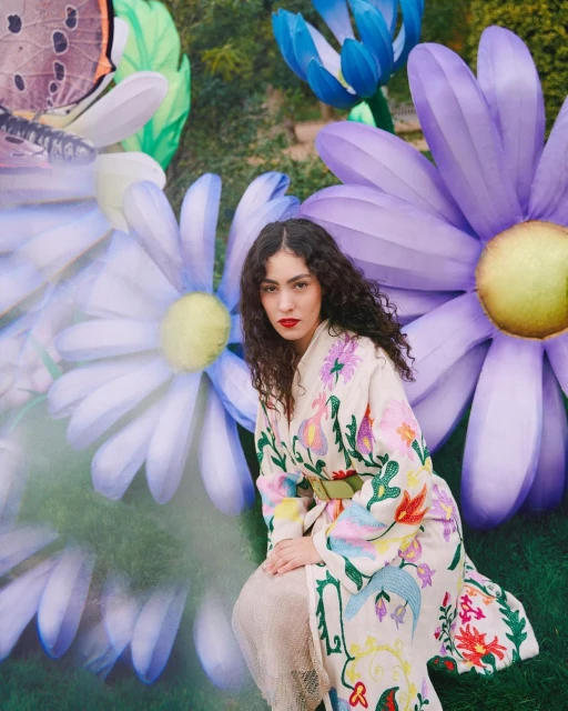 A woman with curly hair kneels in a garden setting with large floral and butterfly decorations, wearing a colorful, embroidered coat.