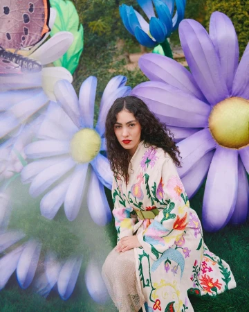 A woman with curly hair kneels in a garden setting with large floral and butterfly decorations, wearing a colorful, embroidered coat.