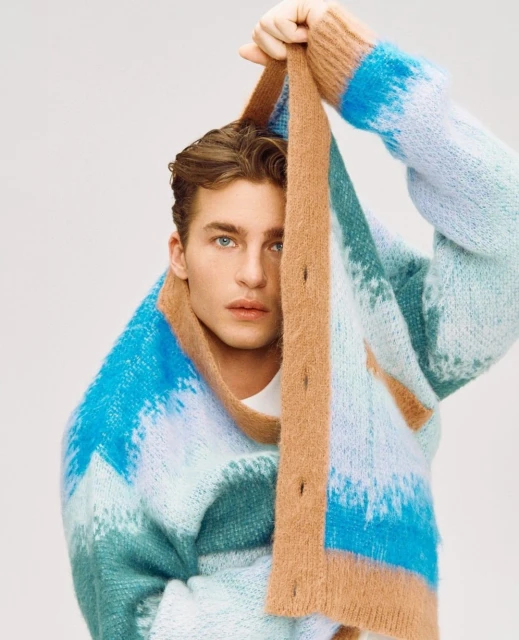 Close-up of a young adult man with blue eyes and brown hair wearing a multicolored striped cardigan in a studio with white background.
