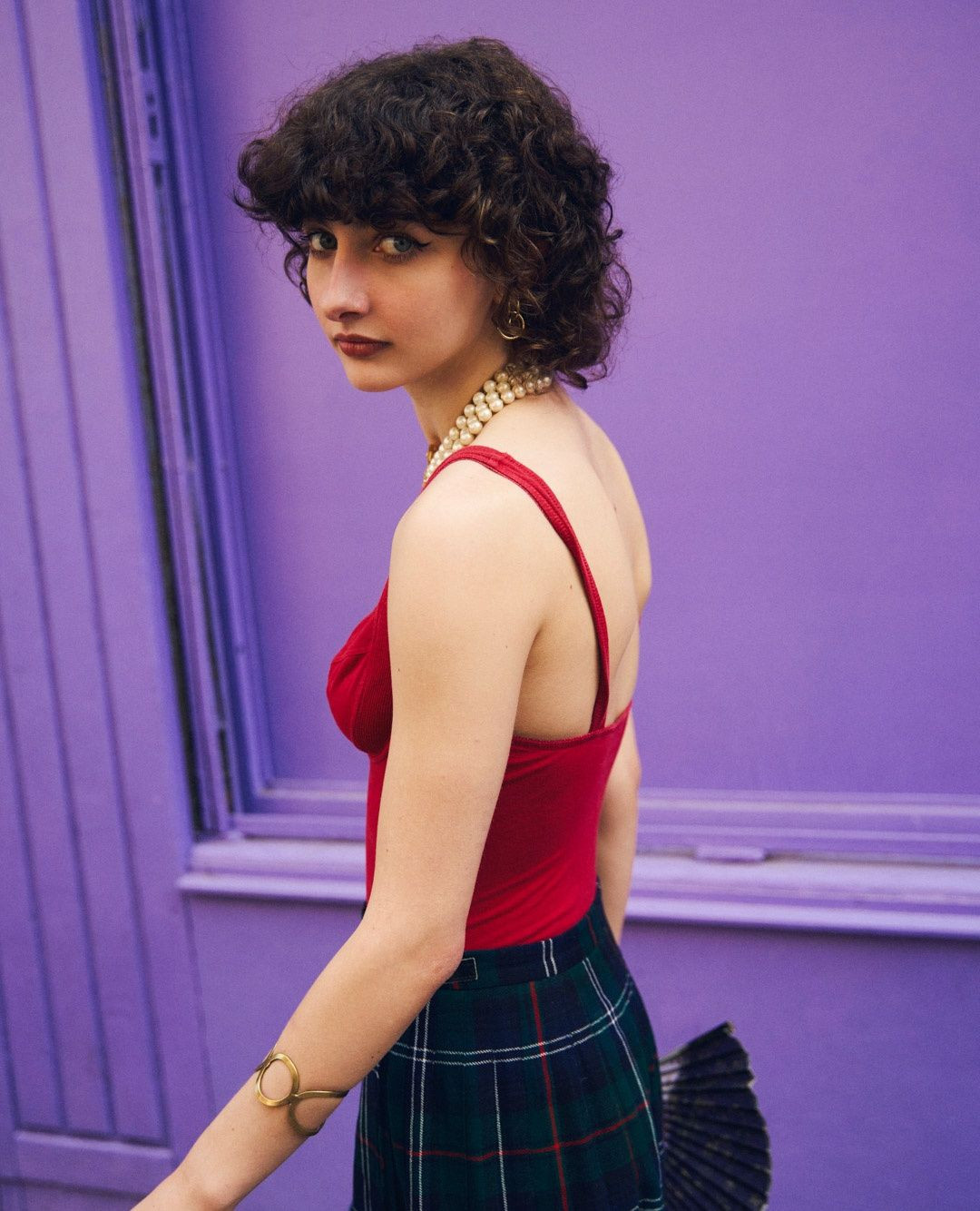 Young woman with curly hair wearing a red top and green plaid skirt stands against a purple wall with a serious expression.