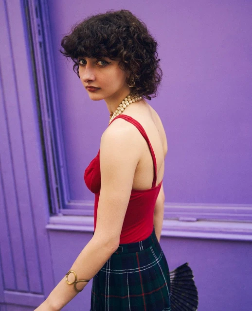 Young woman with curly hair wearing a red top and green plaid skirt stands against a purple wall with a serious expression.