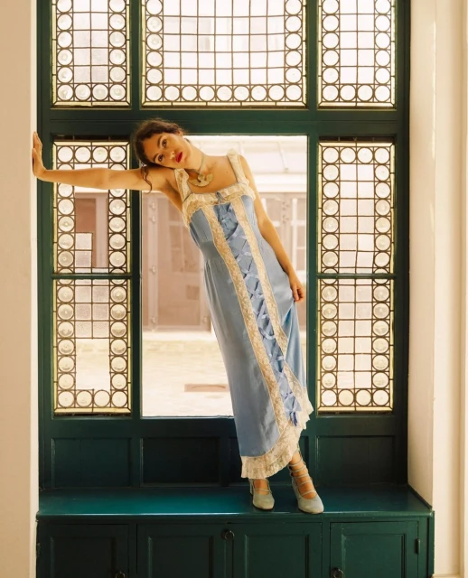 Woman in a blue dress leans against a window with a detailed grid pattern, creating a vintage and elegant fashion portrait.