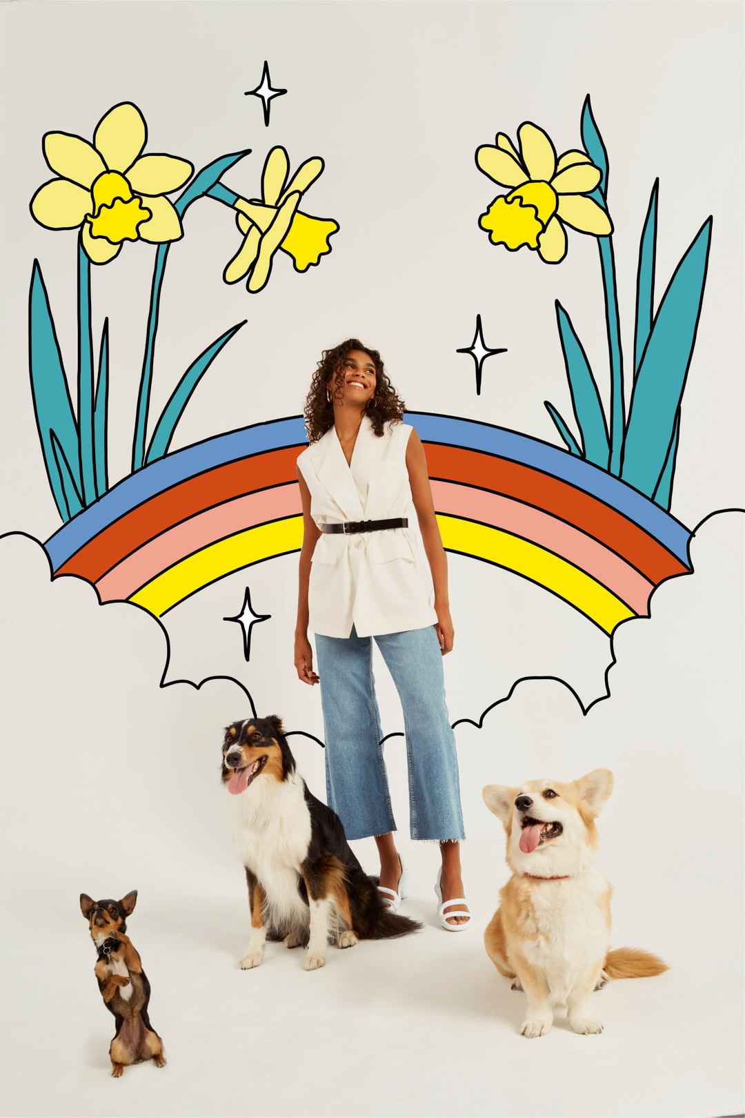Woman with curly hair in a white vest and jeans, posing in a photo studio with a colorful rainbow background and three adorable dogs.