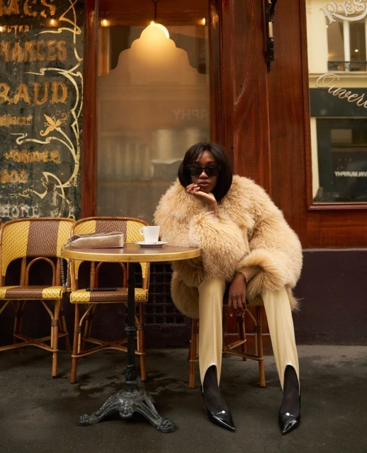 A stylish woman in a fur coat sitting at a Parisian cafe table with a coffee cup, chairs, and ornate building details.