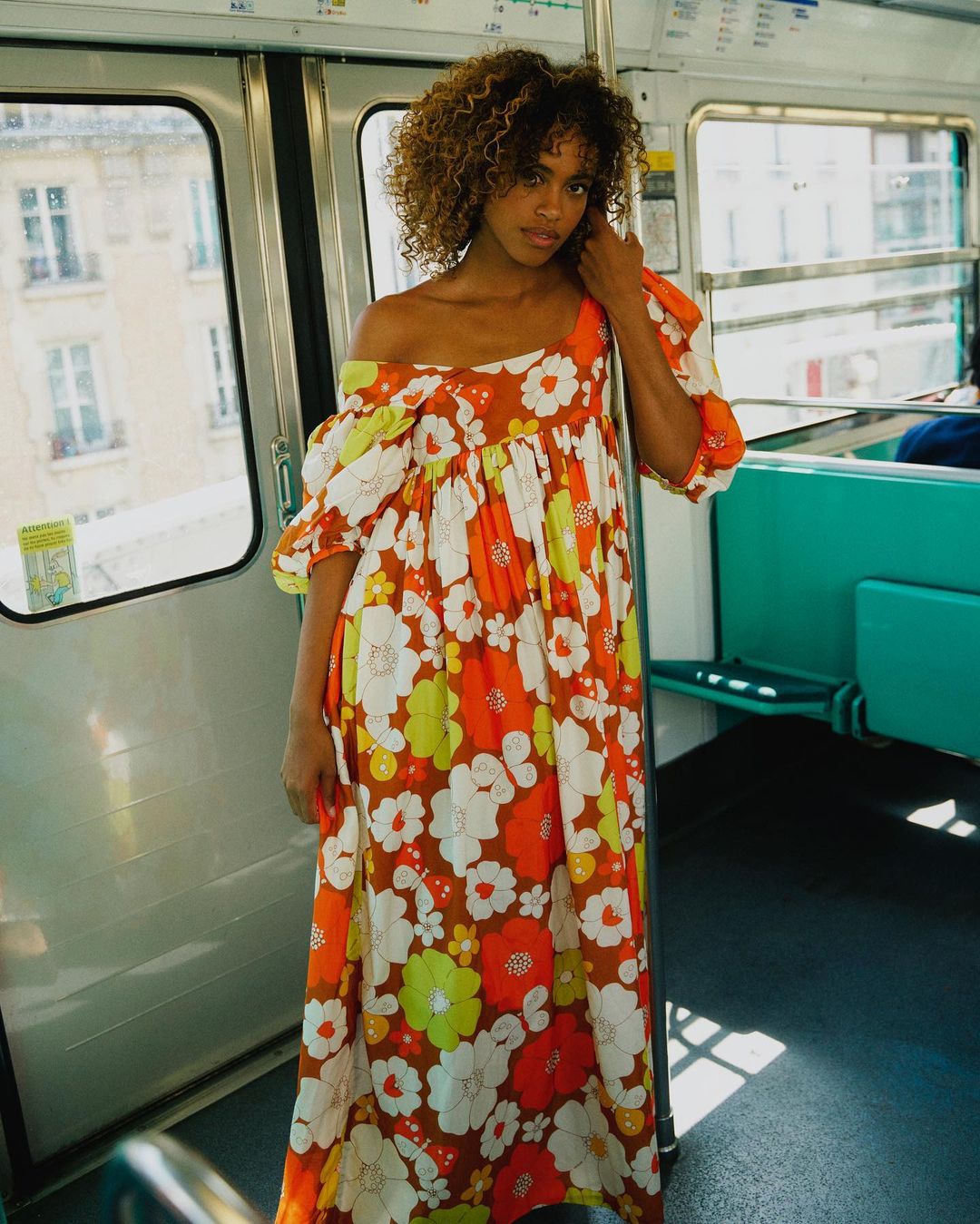 Adult woman with curly hair in floral dress standing inside a train car with teal seats, windows, and white walls, portrait orientation.
