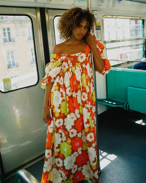 Adult woman with curly hair in floral dress standing inside a train car with teal seats, windows, and white walls, portrait orientation.