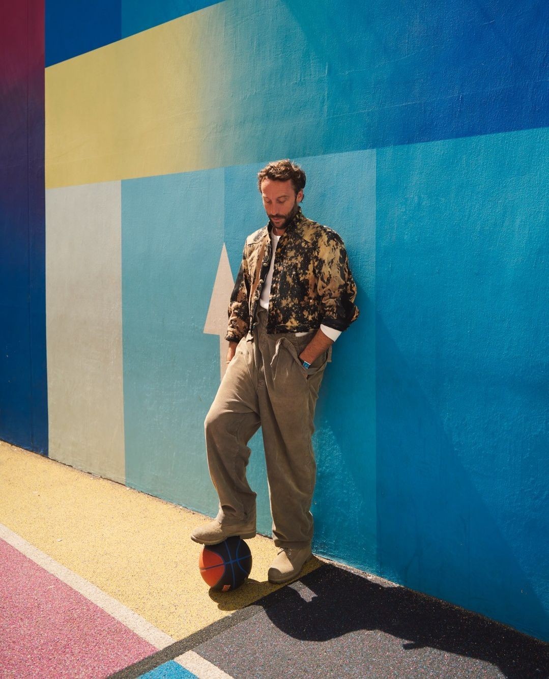 An adult male balances his foot on a basketball near a colorful painted wall, wearing a patterned jacket and casual pants, captured in daylight.
